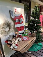 Wide view of the artificial Christmas tree, large wooden Santa decoration, wreath, assorted small decorations on a wooden table.