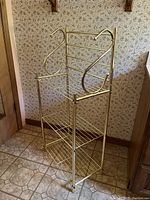 Photo showing a three-tier chrome metal shelf unit with wire shelves and decorative curved metal side railings against a floral wallpaper background.