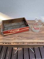 Photo showing wooden Coca-Cola crate and glass pitcher on a wooden surface from the top front angle