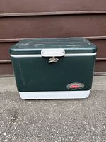 Front view of dark green Coleman steel cooler with white base and latch, placed outdoors on concrete against brown wooden wall.