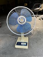 Electric table fan with blue blades and metal protective grill, beige base with control buttons on control panel. Taken to show good physical condition and color detail.