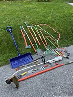 Wide shot of garden tools including rakes, shovels, hand tools, and saws lined up on a driveway with grass background.