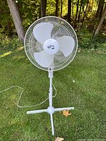 Full view of Hampton Bay white floor fan on grass background showing three blades, circular grill, adjustable stand, and tripod base with power cord visible.