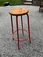 Red metal stool with round wooden seat showing wear and paint marks, standing on concrete surface outdoors.