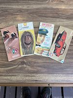 Four vintage road maps with varied cover artwork laid out on a wooden surface; includes wildlife, Native art, law enforcement figure, and vintage automobile.