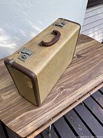 Closed view of the vintage suitcase showing beige exterior, brown leather corner trim and brown handle, and metal clasps.