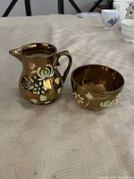Photo of the vintage Wade copper luster ware creamer and cup with floral designs on a textured surface.