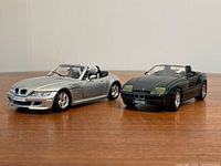 Photo of two BMW die cast model cars side by side on a wooden surface, showing the full body of the silver BMW M Roadster and green BMW Z1 convertible models.