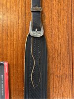 Close-up of black leather guitar strap with silver buckle and embossed decorative patterns.