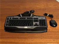 Logitech G15 keyboard and Logitech G9 mouse laid out on a wooden table, showing the full keyboard and mouse connection cables.