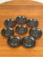 Top down view of 8 dark brown stoneware saucers arranged on a wooden table, showing flat center and round rims.