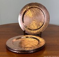 Photo showing five copper charger plates stacked with one plate displayed upright on a wooden table, emphasizing the hammered texture and shiny copper finish with patina.