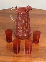 Photo showing cranberry glass pitcher with four matching cups on wooden surface to display overall set condition and size.