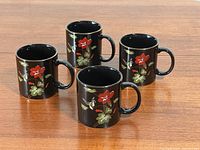 Four black ceramic Japanese mugs with gold-plated rims, red flowers, white birds, and green leaves, arranged on wooden table.