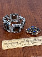 Photo showing silver-tone linked bracelet with square smoky grey stones next to floral mosaic brooch on wooden surface with ruler for scale.