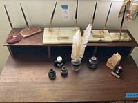 Wide shot showing assortment of desk pens on wooden and marble bases, two white feather quills in glass ink pot, and ink bottles on desk surface.