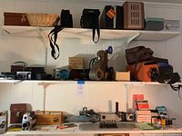 Shelf with multiple vintage cameras in leather pouches, camera cases, a wicker basket, and projector equipment parts scattered across two white shelves against a white paneled wall.