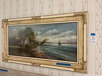 Angled view of framed scenic wall art showing the painting of a cottage by the ocean with a sailboat in the distance.