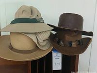 Group of six hats: tan canvas outdoor hat, beige straw hat, and four brown leather western-style hats shown on stands.