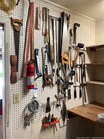 Wide view of pegboard with assorted hand tools hung including saws, hammers, mallets, squares, and other small hand tools.