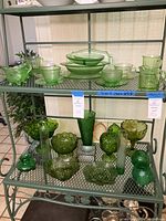Photo showing multiple green depression glass items including footed bowls, vases, and a covered box, arranged on green metal shelving.