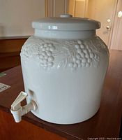 Front and side angle of the white ceramic beverage dispenser showing grapevine motif and plastic spout attached near the base.