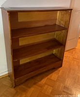 Front and side view of wood bookcase showing three shelves, medium brown stain, unfinished back panel and scalloped base trim.