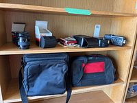 Wide view of a wood shelf displaying two black camera cases on bottom shelf and various boxed camera accessories including flashes and lens items on shelf above.