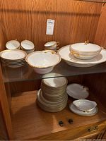 Display of cups and saucers, bowls, plates, covered dish, gravy boat, platter and serving dishes with gold trim in wood cabinet.