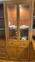 Full front view showing oak bookshelf with glass doors, illuminated top light, drawer, and cabinet base containing white dishware (items not included).