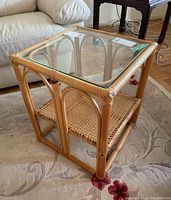 View of the rattan side table showing the clear glass top and woven lower shelf on a carpeted floor near a sofa.
