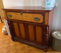 Front angle view of wood sideboard showing birds-eye maple panels, drawer with metal handles, and cupboard doors below along with carved legs and base.