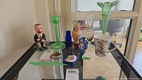 Overview of lot on glass shelf featuring various vases, a pill box, key chain, jug, coffee mug, and figurine