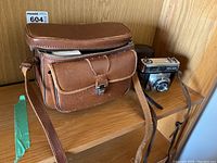 Photo showing the vintage leather carrying case with multiple compartments and shoulder strap beside the Zeiss Ikon camera placed on wooden shelf.