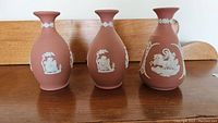 Three Wedgwood jasperware vases, 5 inches tall, terracotta color with white relief designs, sitting on a wooden surface.