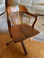 Front and side view of medium-tone wood desk chair with curved arms and vertical slat backrest. Chair sits on four legs with casters, one caster noted as loose.