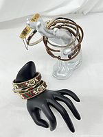 Photo showing multiple bracelets displayed on a silver hand mannequin and black hand mannequin; includes goldtone bangles, brown twisted cable cuff, silver cuff with pyramid accents, and fabric-wrapped bead embellished bangles.