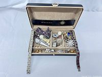 Open small black jewelry box showing vintage earrings, brooches, and bracelets inside