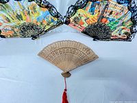 Three vintage hand fans displayed on a flat surface; two large colorful Spanish-style fans with cloth leaves and black lace trim at the back, one smaller carved wooden Chinese fan with a red tassel in front.