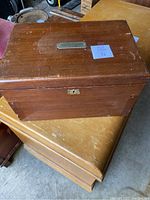 Closed view of the antique wooden sewing kit box placed on top of wooden furniture, showing front and top surfaces with metal nameplate and lock keyhole.