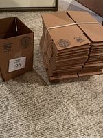 View showing one box assembled and one stack of 24 flat boxes tied with a rubber band on carpet.