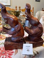 Two carved wooden elephant bookends shown from front angle on table with floral cloth and various silver dishware in the background.