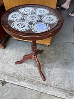 Full view of round wooden accent table with glass top and lace doily inside, showing tripod base and scratches on wood.
