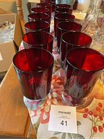 Front view showing full set of 12 ruby red glasses in rows on wooden table with floral tablecloth.