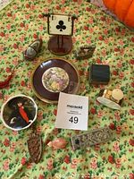 Photo of assorted miniatures and decorative odds arranged on floral tablecloth including plates, figurines, card stand, and other small items.