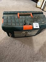 Closed black plastic tackle box with orange details and latches, placed on carpeted floor