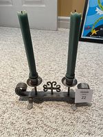 Full front view of the pewter candelabra holding two green taper candles placed on a carpeted floor with a framed poster in the background.