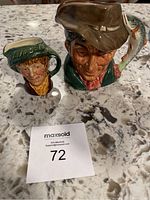 Two miniature Royal Doulton Toby jugs showing detailed faces and varying hat styles on a marble surface next to a MaxSold number card.
