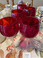 Photo showing five ruby red wine glasses with twisted clear glass stems on floral tablecloth with various decorative items in background.