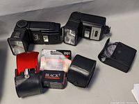 Two black camera flashes and three camera accessories including a flash diffuser, teleconverter, and box of flashbulb